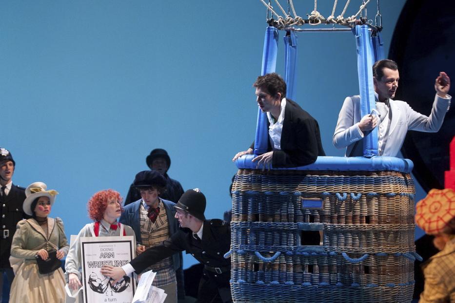 Eine Theaterszene mit Menschen in Kostümen, einem Heißluftballon und einer Telefonzelle.