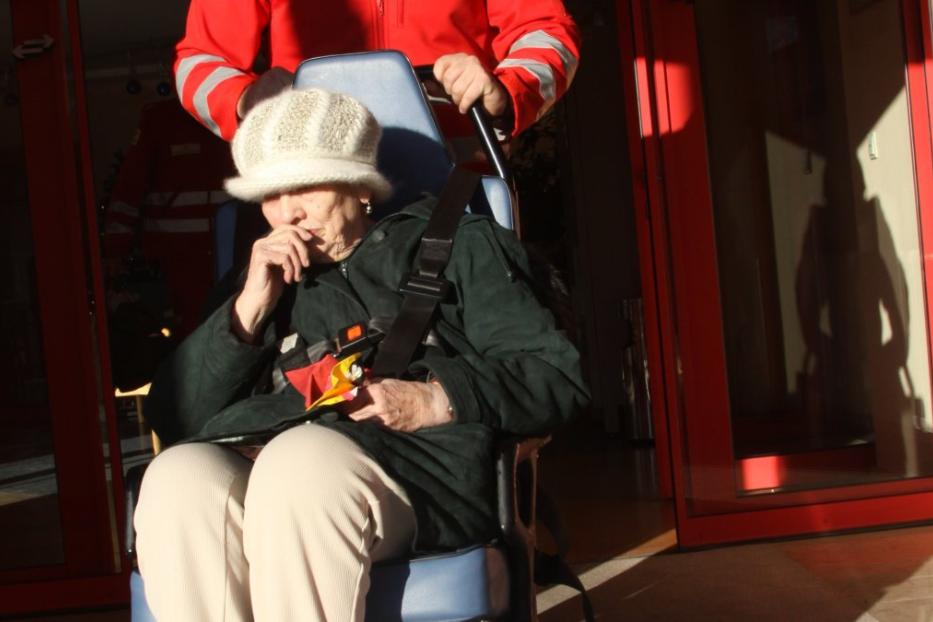 Eine ältere Frau sitzt gesichert in einem Rollstuhl, der von einem Sanitäter geschoben wird.