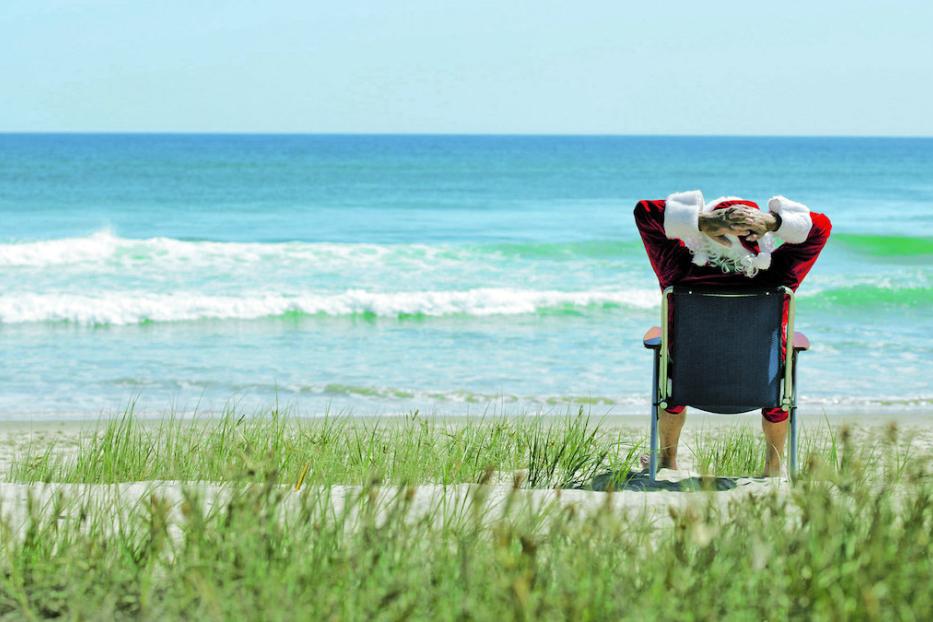 Ein Weihnachtsmann entspannt im Liegestuhl am Strand und blickt auf das Meer.