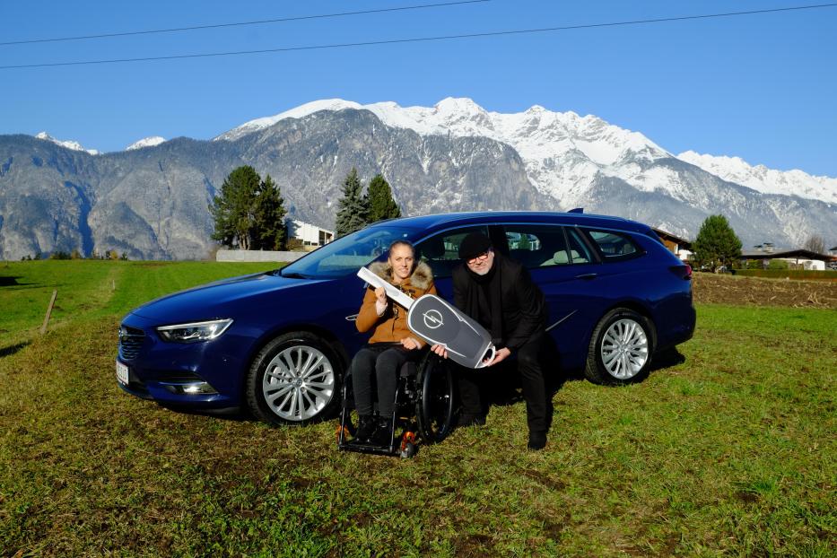 Eine Frau im Rollstuhl und ein Mann präsentieren einen übergroßen Opel-Schlüssel vor einem Auto und Bergpanorama.