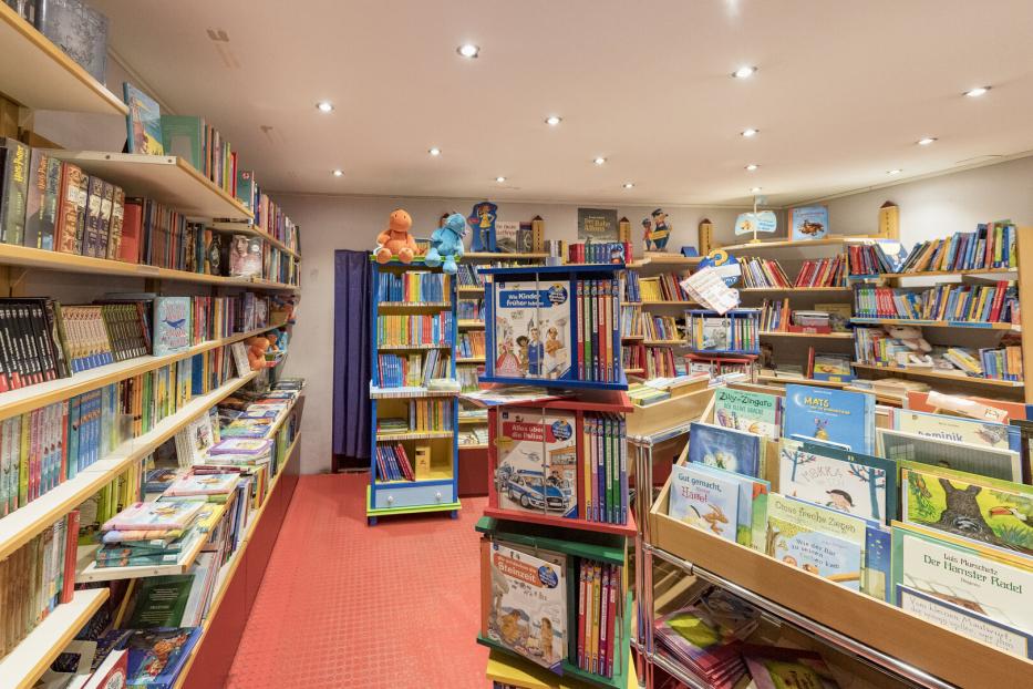 Regale voller bunter Kinderbücher, einige Stofftiere sitzen auf den oberen Regalen, helle Beleuchtung und roter Boden.