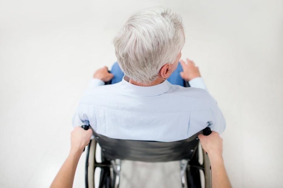 Ein älterer Mann mit grauen Haaren sitzt im Rollstuhl und wird von einer Person von hinten geschoben.