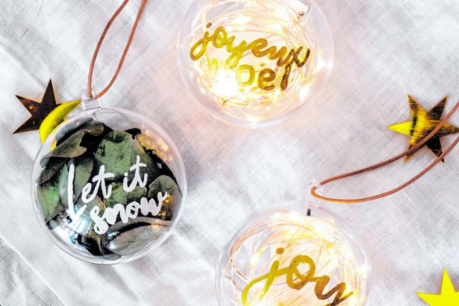 Drei transparente Weihnachtskugeln mit den Aufschriften „Joyeux Noel“, „Let it snow“ und „Joy“.