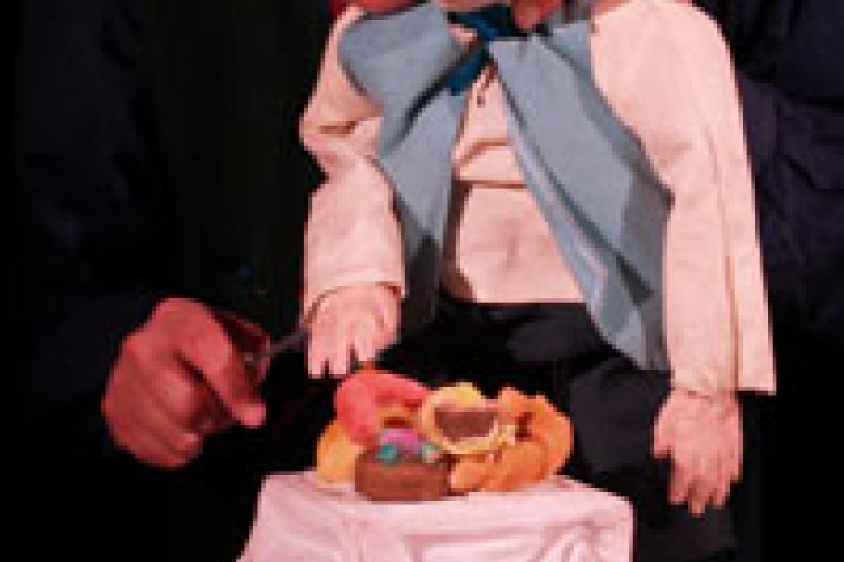 Ein Mann mit Brille und roter Mütze spielt mit einer Marionette an einem Tisch mit Essen.