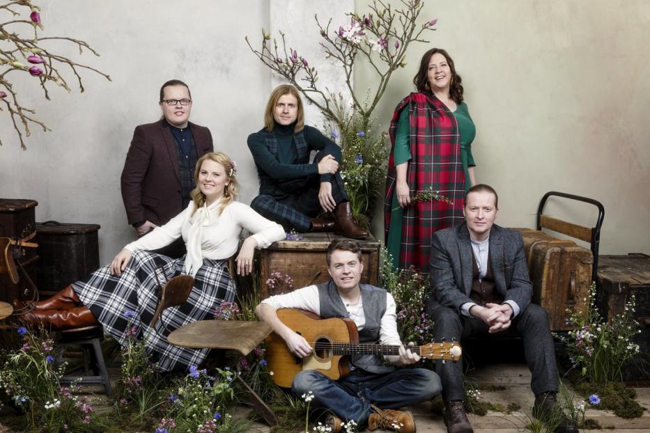 Eine Gruppe von sechs Personen posiert inmitten von Blumen und alten Koffern, einer spielt Gitarre.