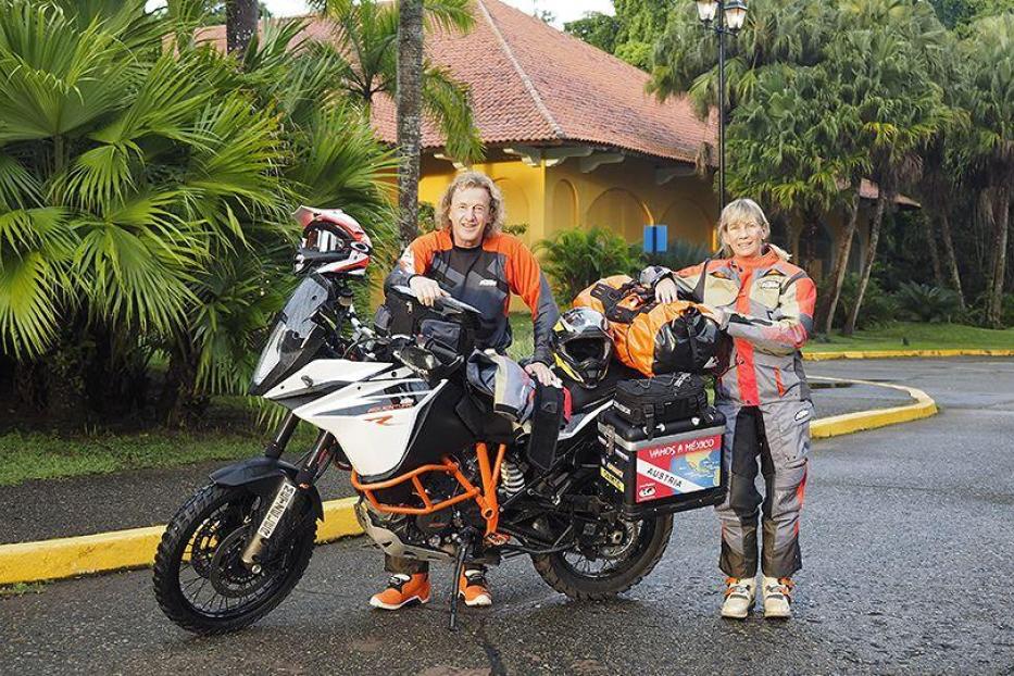 Zwei Motorradfahrer, ein Mann und eine Frau, posieren mit ihrem vollgepackten Motorrad vor Palmen.