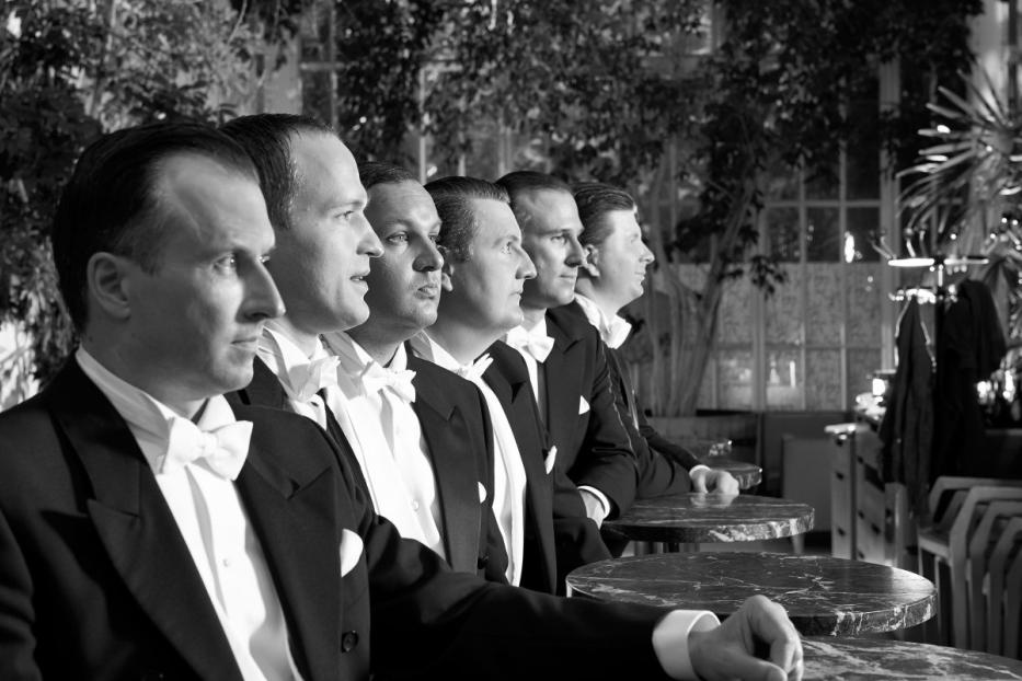 Die Wiener Comedian Harmonists in Anzügen und Fliegen stehen an Tischen.