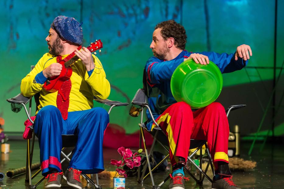 Zwei Clowns sitzen auf Stühlen, einer spielt Ukulele, der andere hält eine grüne Schüssel.