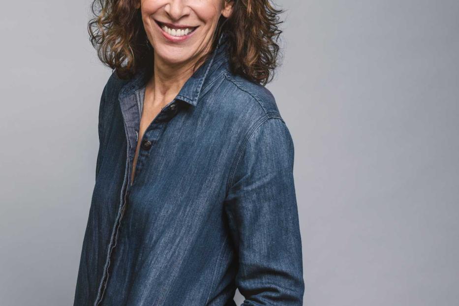 Adele Neuhauser lächelt in einem blauen Jeanshemd vor einem grauen Hintergrund.