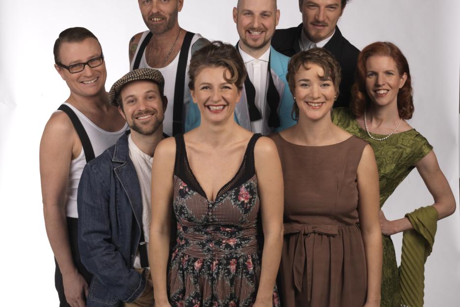 Eine Gruppe von acht Personen in Kostümen posiert lächelnd vor einem weißen Hintergrund.