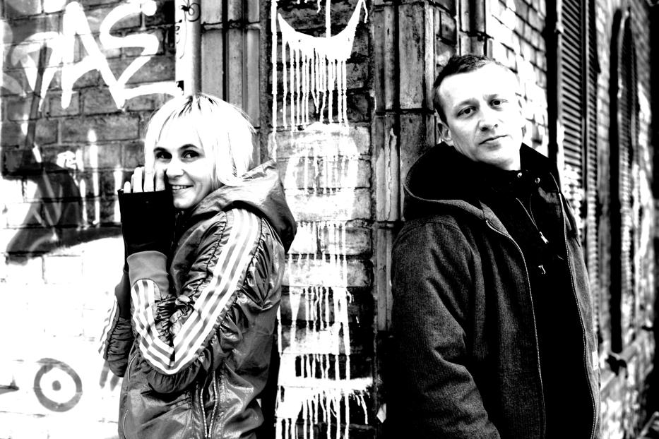 Schwarzweißfoto von zwei Personen vor einer Wand mit Graffiti.