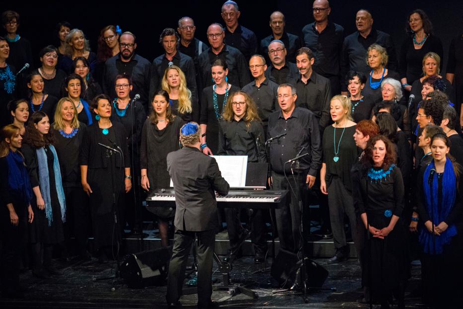 Ein Chor singt auf einer Bühne, begleitet von einem Dirigenten am Klavier.