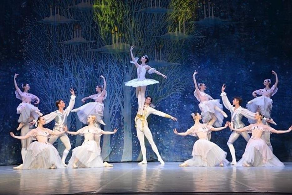 Eine Ballettaufführung mit Tänzern in weißen Kostümen vor einem Bühnenbild mit Bäumen.