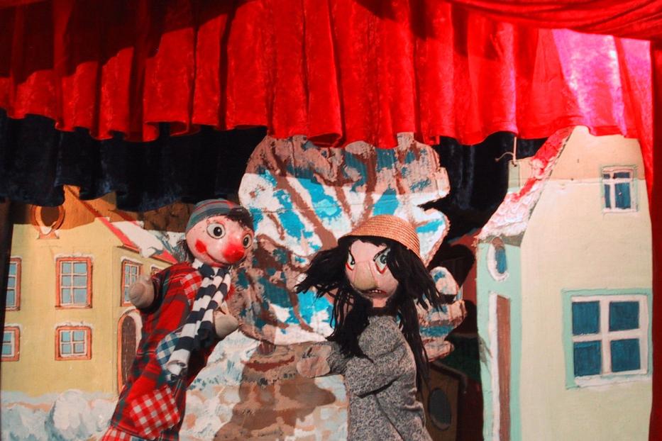 Kasperl und eine Hexe stehen auf einer Puppentheaterbühne vor bemalten Häusern.