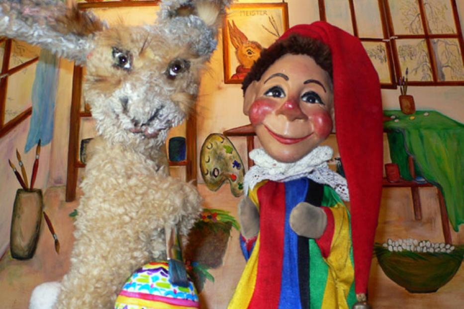 Ein Stoffhase mit einem bemalten Osterei und eine Kasperlepuppe vor einem bemalten Hintergrund.