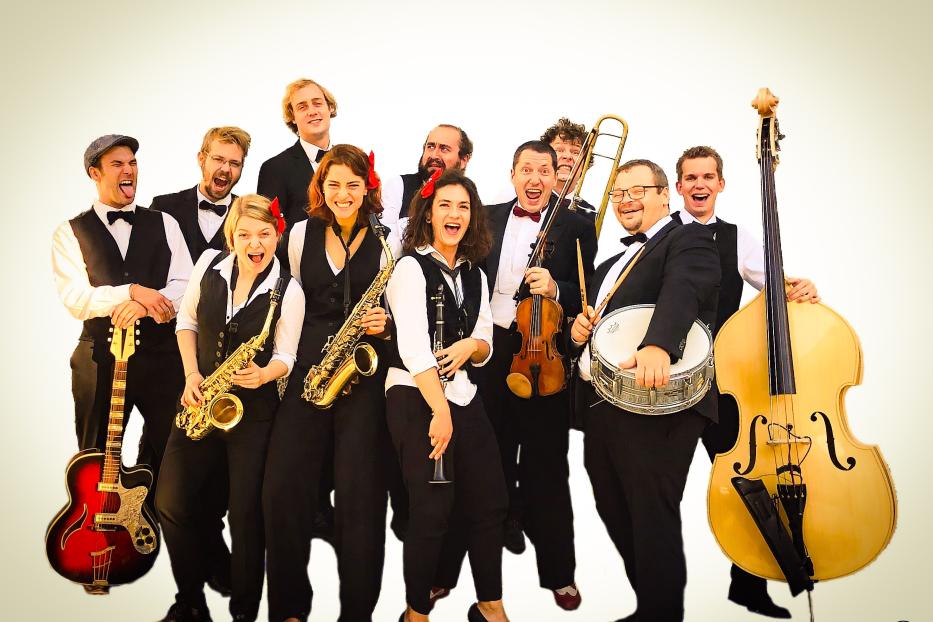 Eine Band mit Blasinstrumenten, Gitarre, Geige, Schlagzeug und Kontrabass tritt auf.