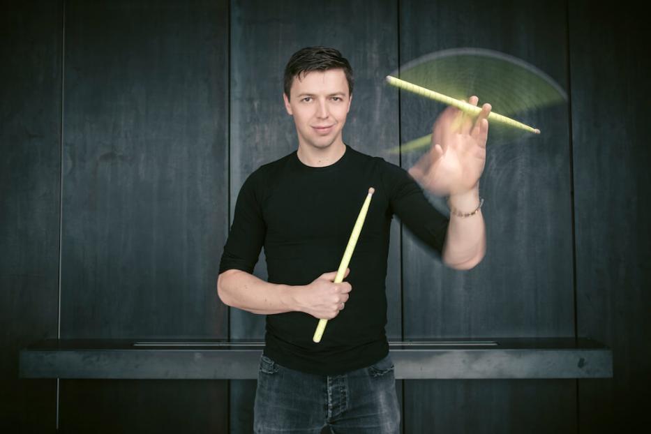 Martin Grubinger jongliert mit Drumsticks vor einer dunklen Wand.