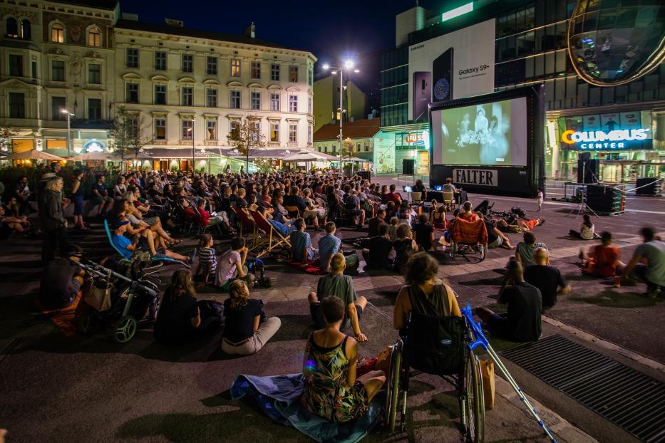 Viele Menschen sitzen im Freien und schauen einen Film auf einer Leinwand an.