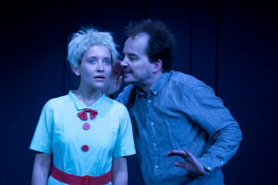 Ein Mann flüstert einer Frau mit blonden Haaren etwas ins Ohr, möglicherweise auf einer Theaterbühne.
