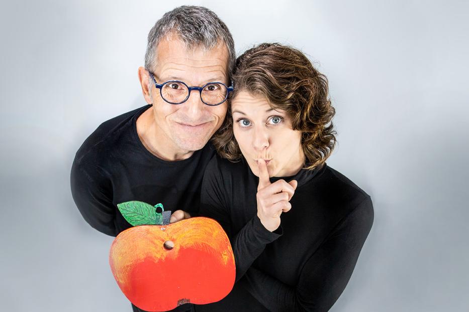 Ein Mann mit Brille hält einen Apfel, während eine Frau den Finger an die Lippen legt.
