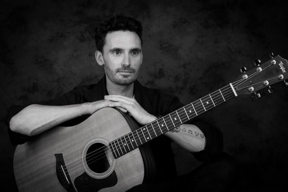 Ein Mann, James Cottriall, posiert mit einer Taylor-Gitarre vor einem dunklen Hintergrund.