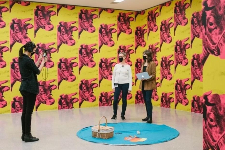 Drei Frauen in einem Raum mit Kuhmotiven von Andy Warhol an den Wänden und einem Picknick auf dem Boden.