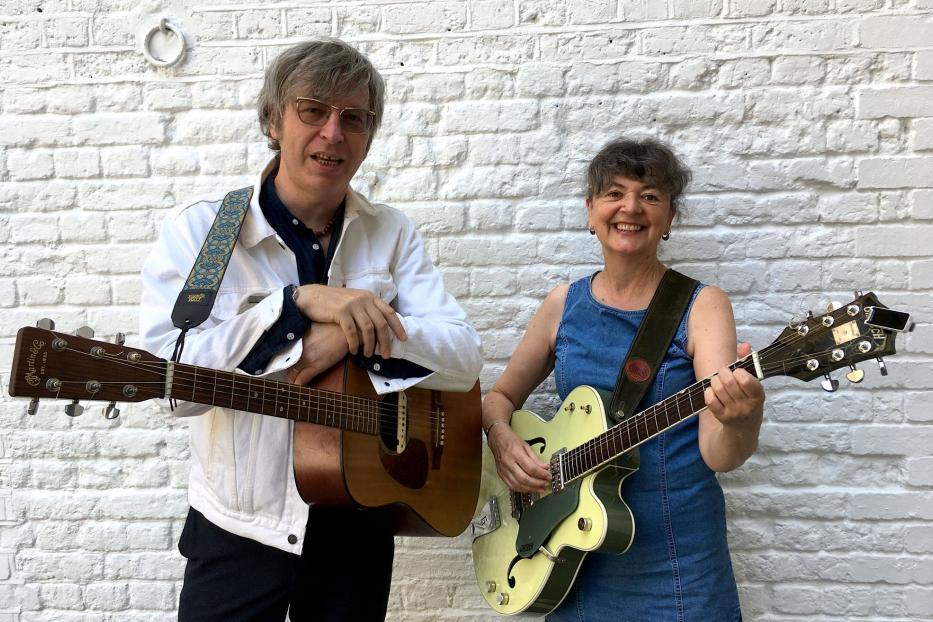 Robert Rotifer und Helen McCookerybook posieren mit Gitarren vor einer weißen Backsteinmauer.