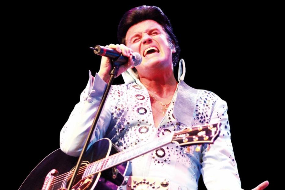 Ein Elvis-Imitator singt mit Gitarre auf einer Bühne.