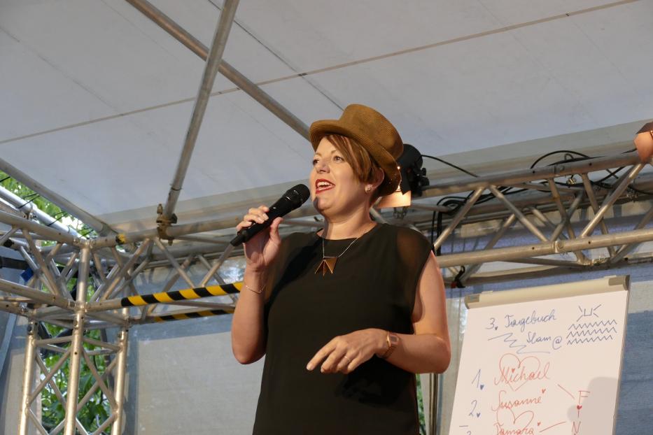 Diana mit Hut moderiert auf einer Bühne mit Flipchart.