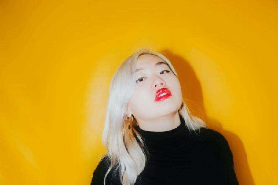 Eine Frau mit blonden Haaren und rotem Lippenstift vor einem gelben Hintergrund.