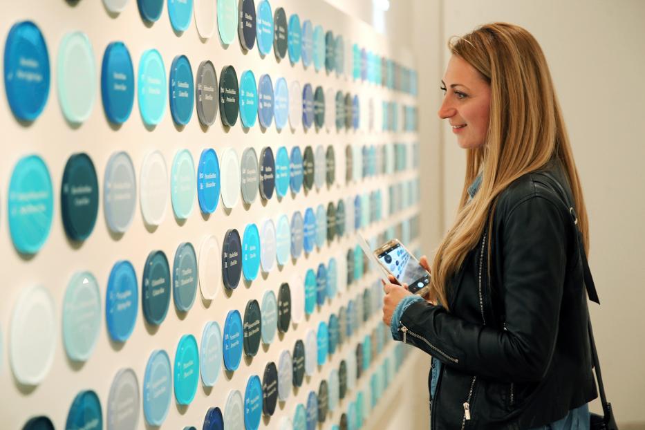 Eine Frau betrachtet eine Wand mit Farbmustern und macht ein Foto mit ihrem Handy.