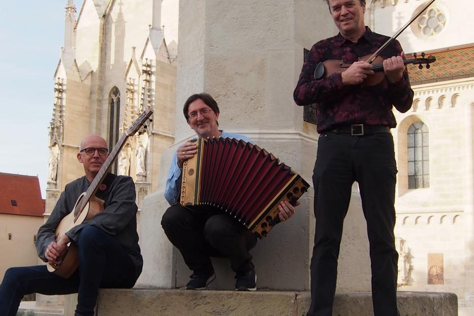 Drei Musiker posieren mit Gitarre, Akkordeon und Geige vor einer Kirche.