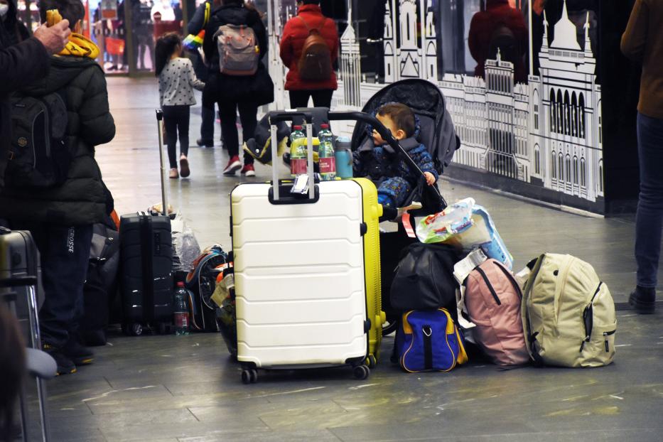 Ein Kind sitzt in einem Kinderwagen, umgeben von Gepäckstücken in einer Wartehalle.