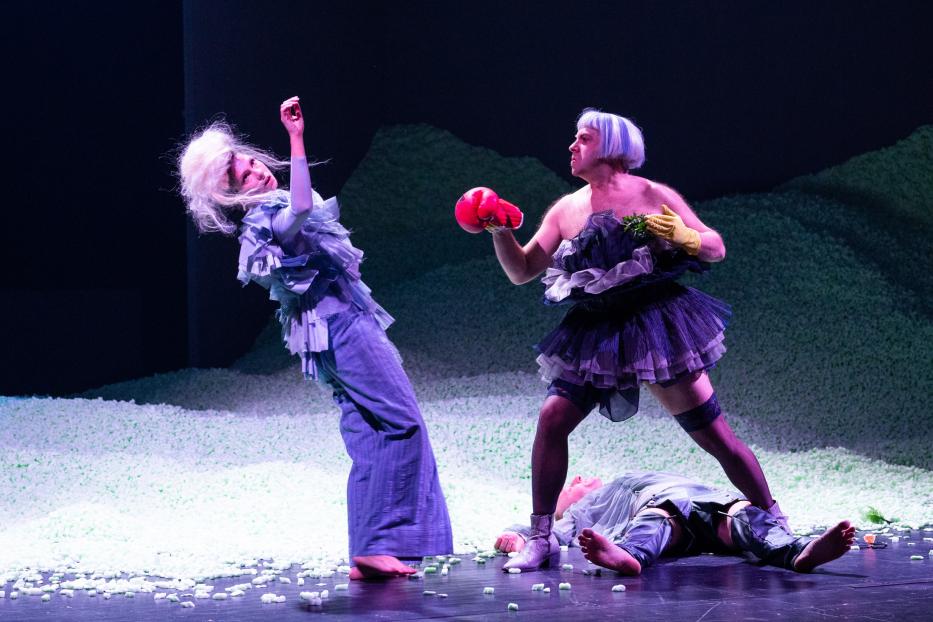 Eine Theaterszene mit drei Darstellern in extravaganten Kostümen auf einer Bühne voller Styropor.