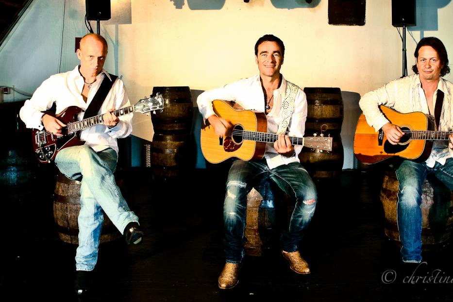 Drei Musiker sitzen auf Fässern und spielen Gitarre.
