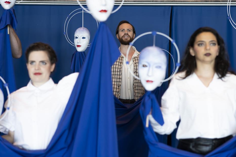 Eine Performance mit drei Personen und schwebenden, weißen Masken vor blauem Hintergrund.