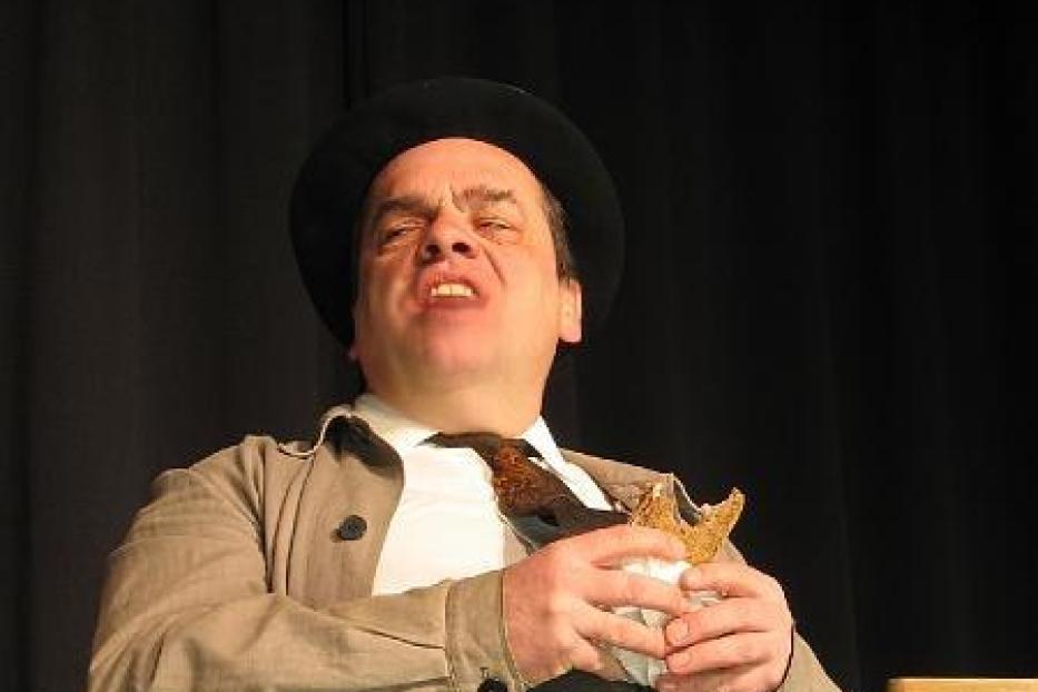 Ein Mann mit Hut und Trenchcoat isst ein Sandwich vor einem schwarzen Vorhang.