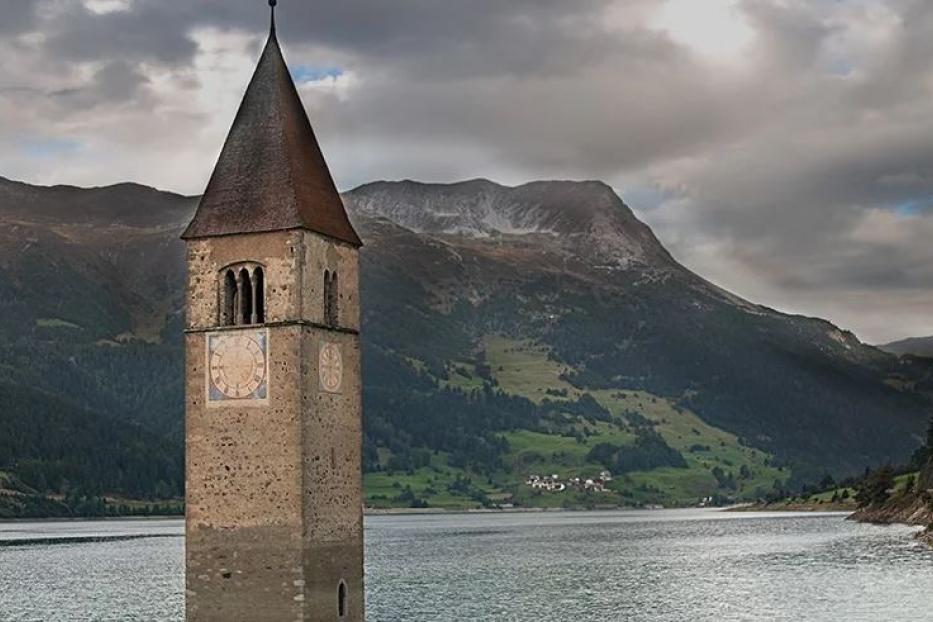 Der Kirchturm von Graun ragt aus dem Reschensee vor einer Bergkulisse.