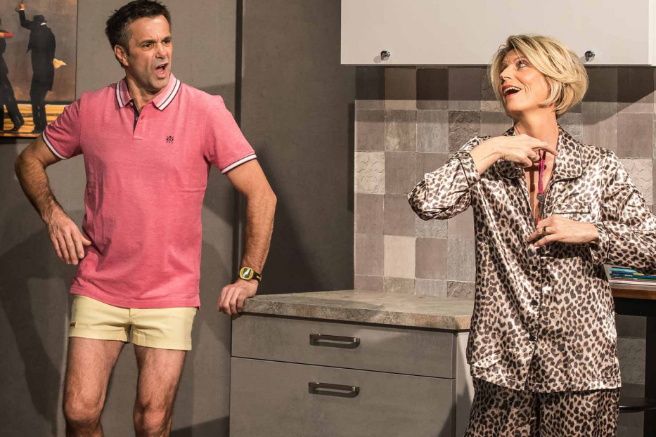 Ein Mann im rosa Polohemd und eine Frau im Leopardenpyjama stehen in einer Küche.