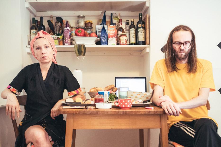 Eine Frau mit Turban und ein Mann sitzen an einem Tisch mit Speisen und einem Laptop.