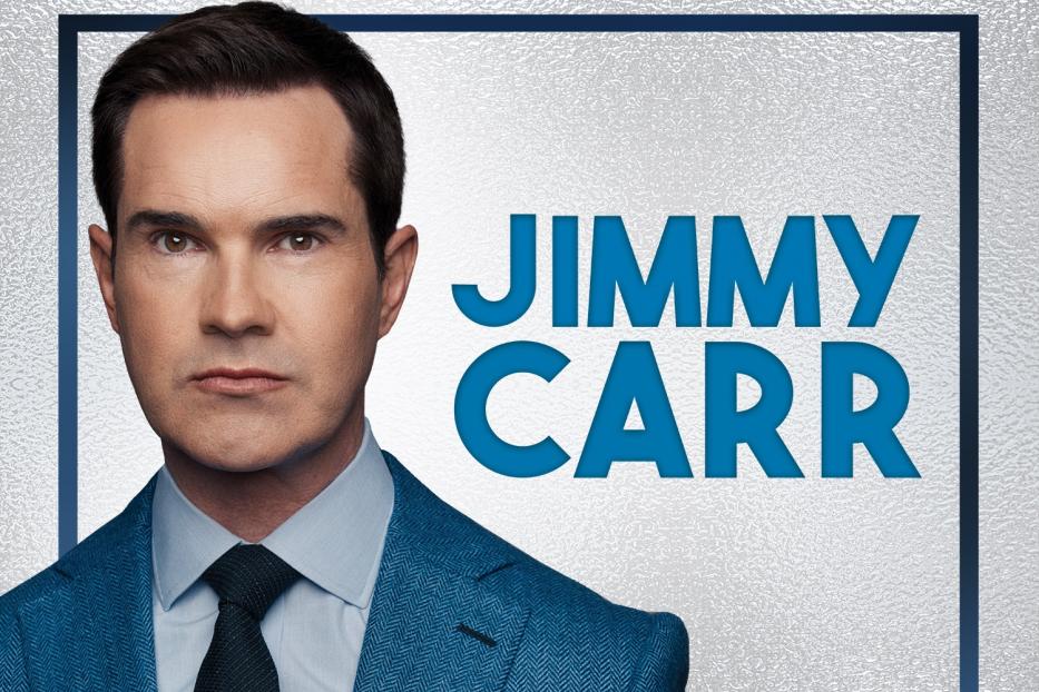 Porträt von Jimmy Carr in einem blauen Anzug vor einem silbernen Hintergrund.