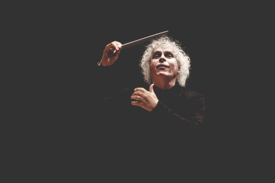 Ein Dirigent mit weißem, lockigem Haar dirigiert ein Orchester.