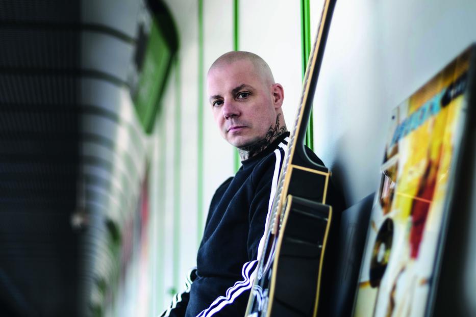 Ein Mann mit Tätowierungen sitzt mit einer Gitarre in einer Unterführung.
