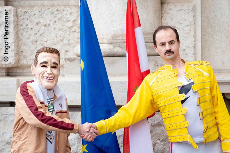 Eine Person mit Arnold Schwarzenegger Maske schüttelt eine Person im Freddie Mercury Kostüm vor EU- und Österreich-Flaggen die Hand.