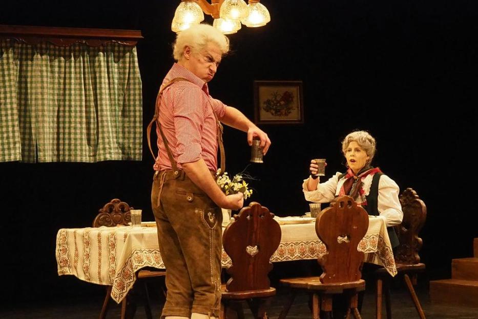 Zwei Schauspieler in Tracht auf einer Theaterbühne an einem Tisch.
