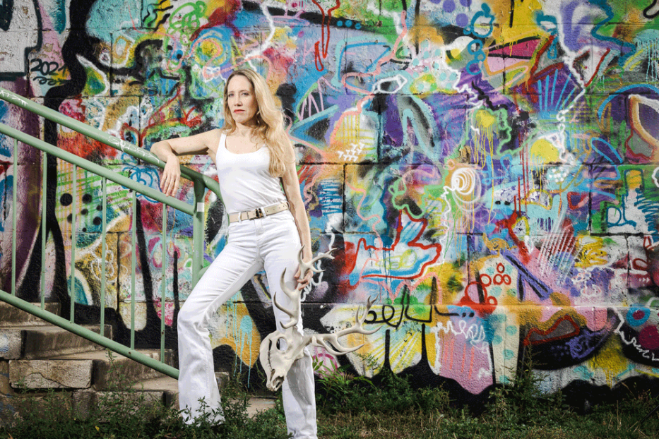 Eine Frau in Weiß posiert mit einem Geweih vor einer Graffiti-Wand.