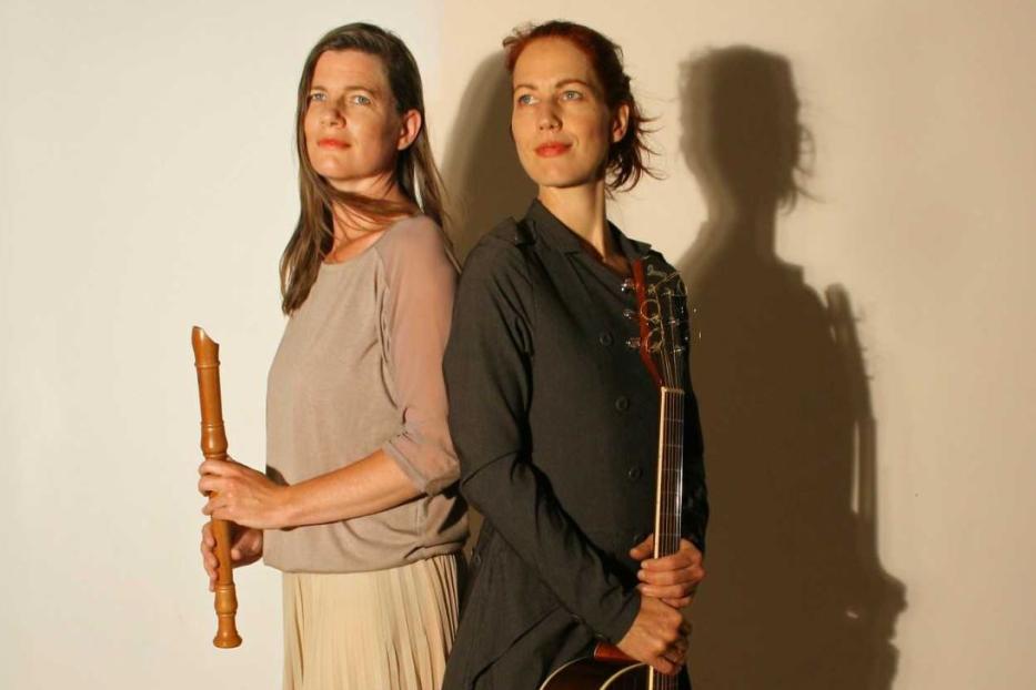 Zwei Frauen stehen Rücken an Rücken, eine mit einer Blockflöte, die andere mit einer Gitarre.