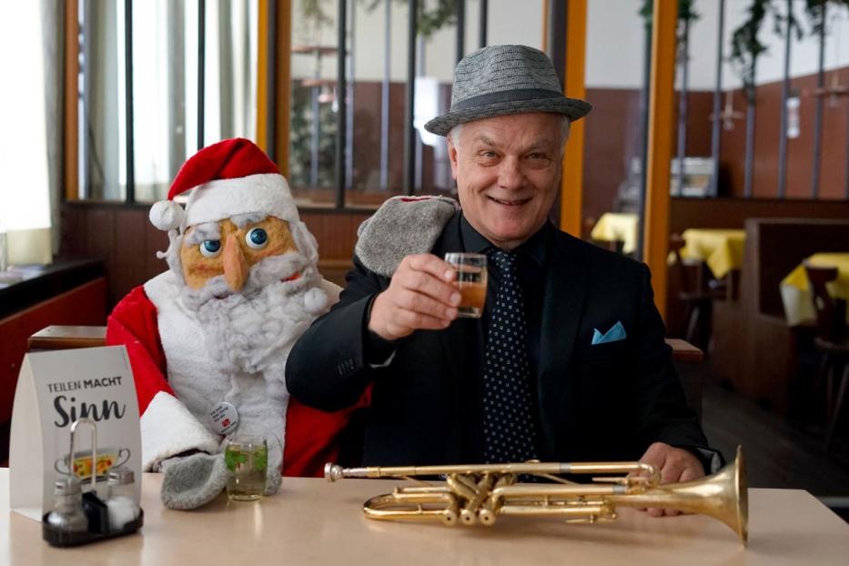 Ein Mann mit Hut prostet neben einer Weihnachtsmann-Puppe mit einem Getränk zu, eine Trompete liegt auf dem Tisch.