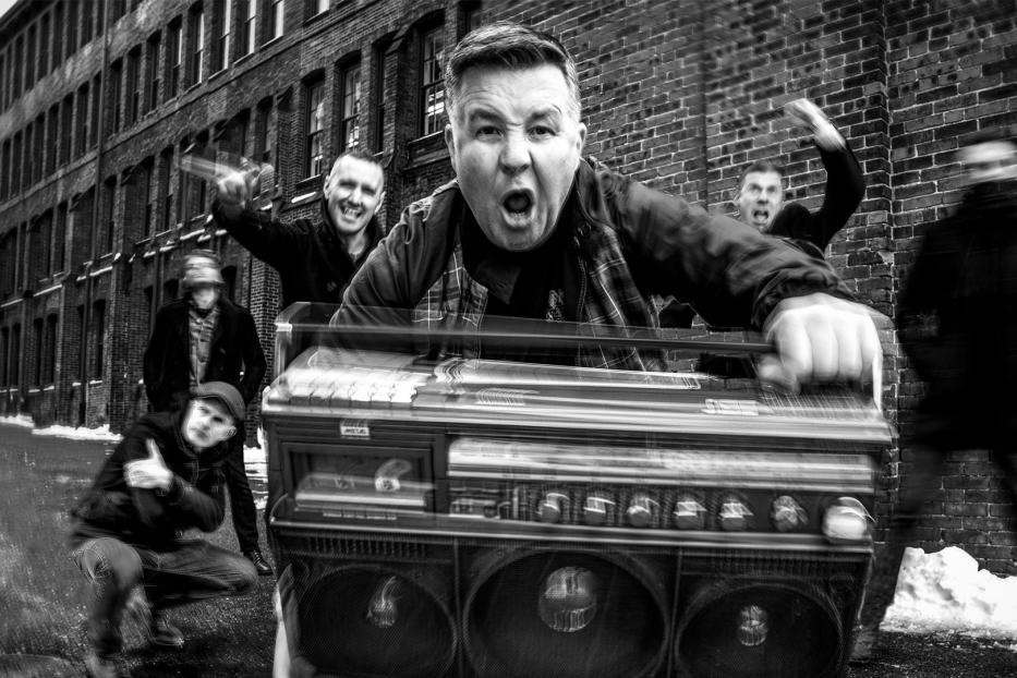 Eine Gruppe von Männern posiert mit einem Ghettoblaster vor einer Backsteinmauer.
