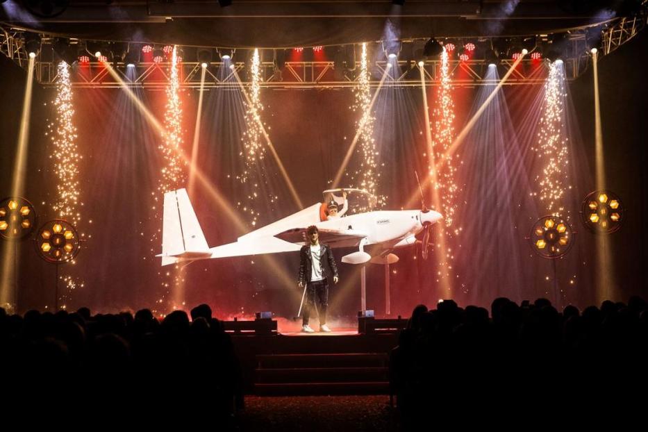 Ein Mann steht vor einem kleinen Flugzeug auf einer Bühne mit Lichteffekten und Publikum.